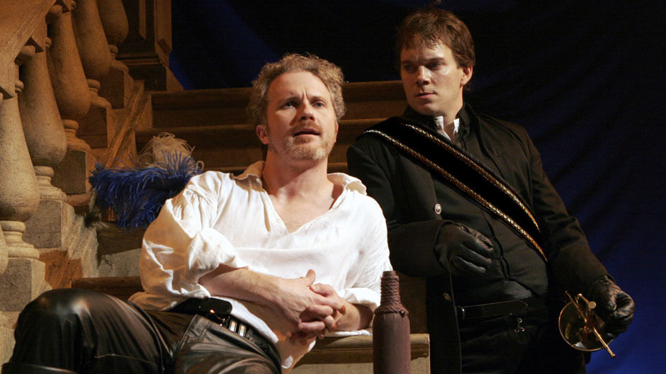 Two men in period costumes on stage; one sits on stone steps in a white shirt and leather pants, looking thoughtful, while the other stands behind him in black attire, holding a sword and gazing intently.