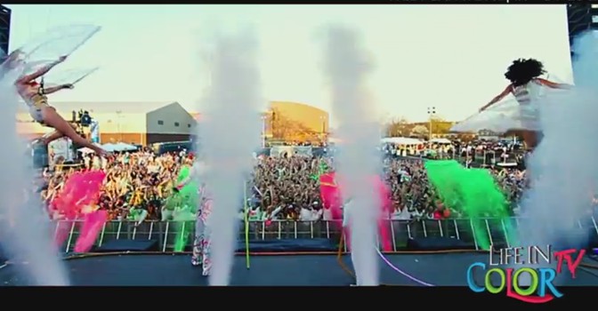 Performers in colorful costumes jump onstage as white smoke and vibrant powder shoot into the air before a large, energetic crowd at an outdoor Life in Color music festival event.