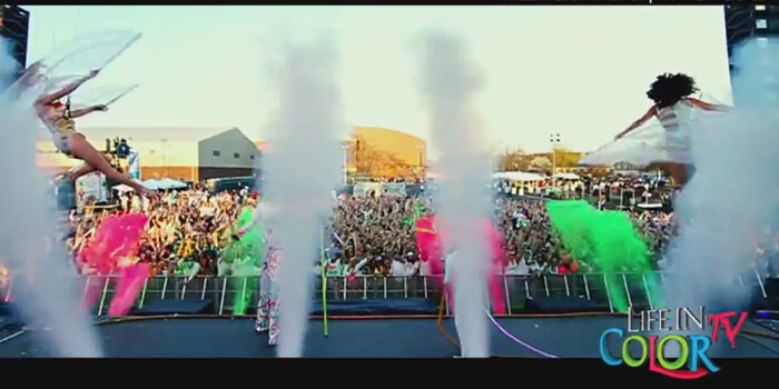 Performers in colorful costumes jump onstage as white smoke and vibrant powder shoot into the air before a large, energetic crowd at an outdoor Life in Color music festival event.