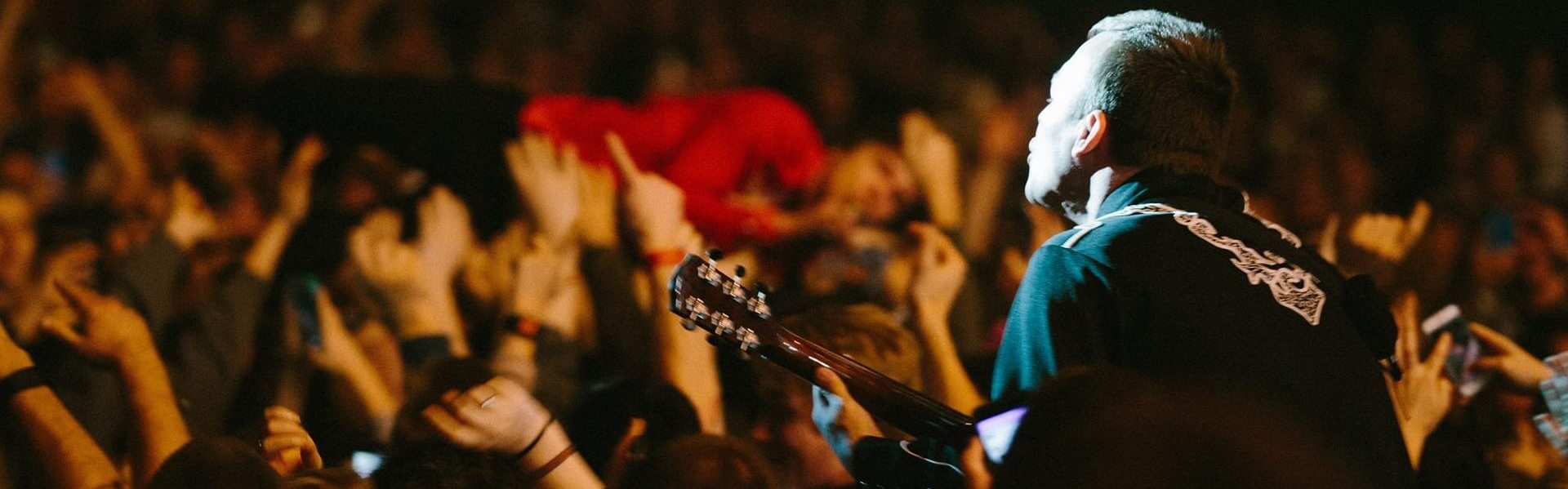 A musician from Cage the Elephant with a guitar performs on stage facing a lively crowd, with many audience members raising their hands and a person crowd-surfing above them.