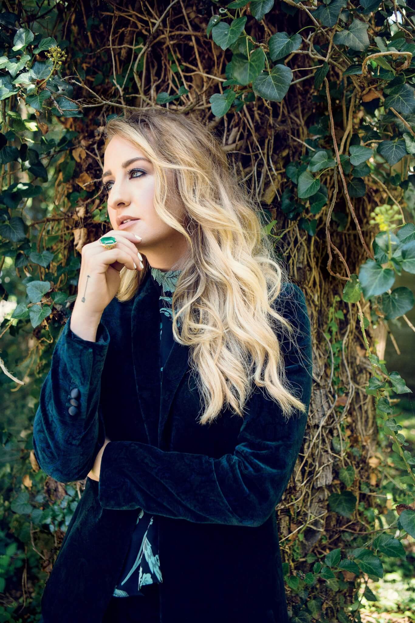 Musician Margo Price with long, wavy blonde hair stands in front of a tree covered in green ivy, wearing a dark velvet jacket and a green ring—posed thoughtfully like Margo Price, a true diamond in the rough. photo by DAVID MCCLISTER