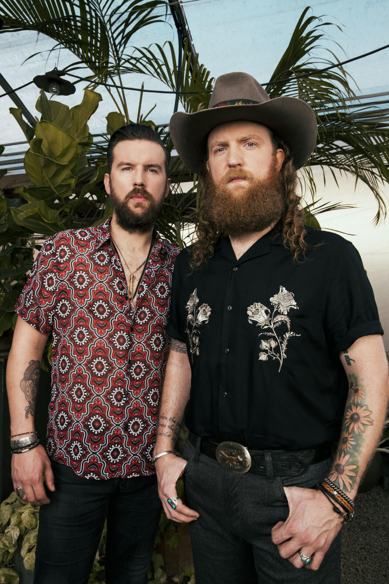 Two men with beards stand side by side in front of green plants, showcasing their unique style for the Alexa Intern Profiles. One wears a patterned red shirt; the other sports a black floral shirt, wide-brimmed hat, and bracelets. Both look toward the camera.