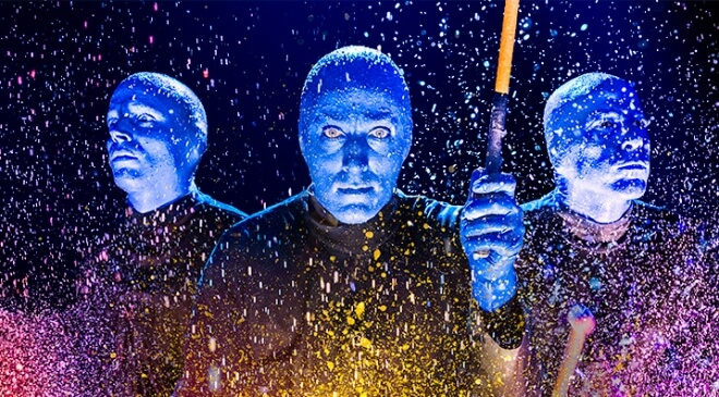 Three performers with painted blue faces and black clothing stand amidst splashes of colorful paint, illuminated by vibrant lighting, creating a dramatic and artistic scene—perfect for Meet Alexa Intern Profiles.