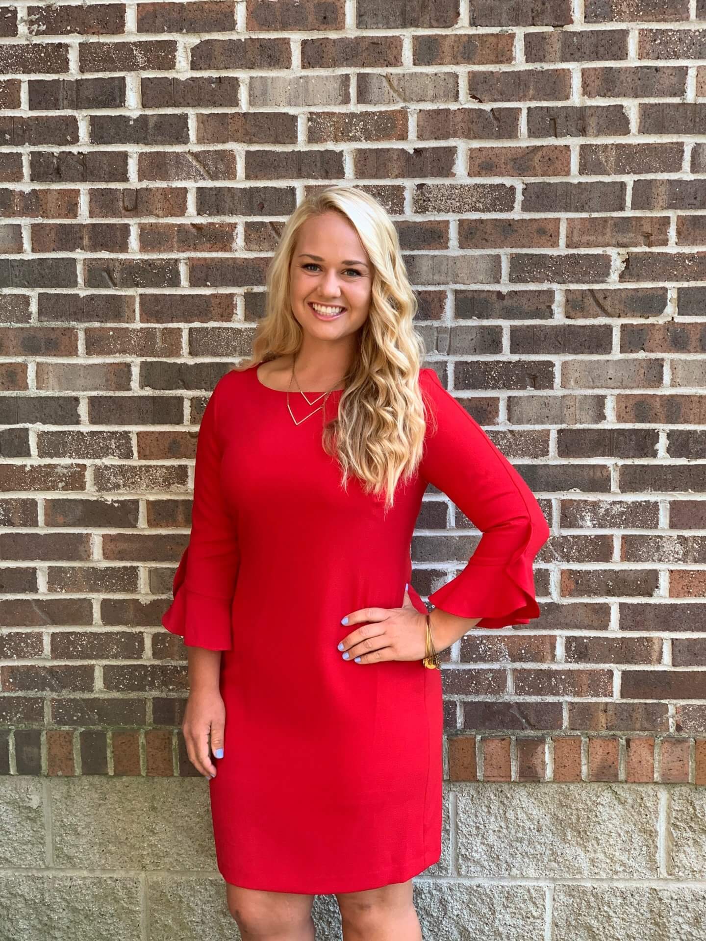 Alexa, a woman with long blonde hair, wearing a knee-length red dress with ruffled sleeves, stands smiling in front of a brick wall with one hand on her hip for the Intern Profiles feature.