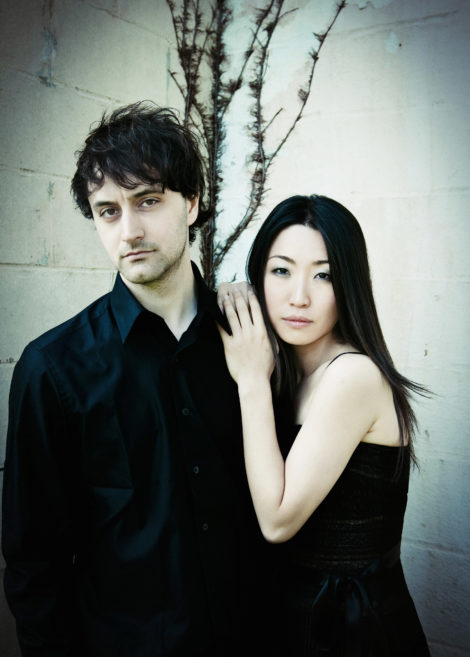 Pianists Ran Dank and Soyeon Kate Lee stand closely together against a pale, textured wall with a bare branch behind them—a striking image from Close Encounters With Music. Both wear black, the woman gently resting her hand on his shoulder.