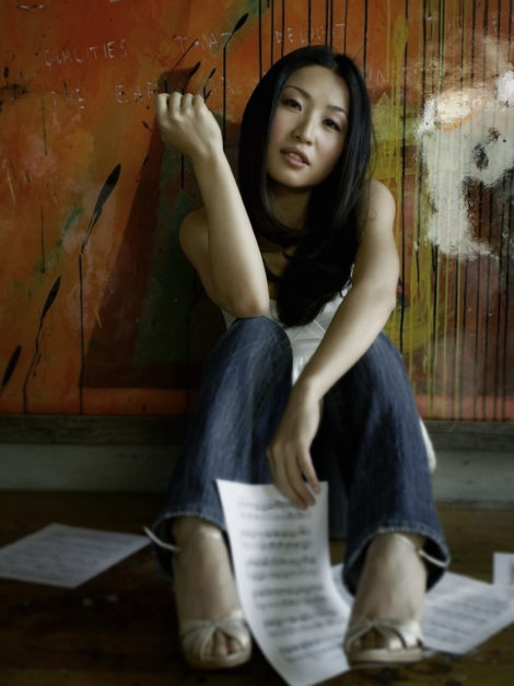 Kate Lee, with long dark hair, sits on the floor in front of an abstract painted wall, wearing jeans and sandals, sheet music from Close Encounters With Music scattered around her. She gazes softly at the camera, one arm resting on her knee.