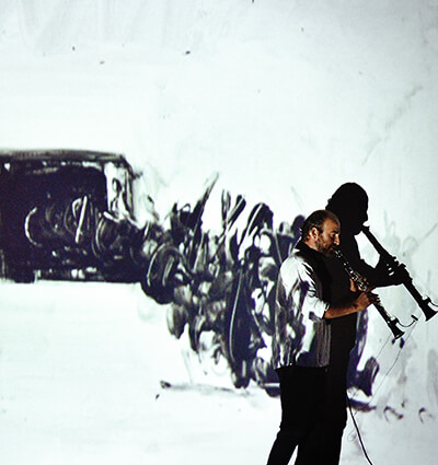 A musician plays a clarinet on stage at the Purdue Convocations Innovative Performance Initiative, with a projected backdrop of stylized, glowing blue figures with raised hands, creating a dramatic and artistic atmosphere.