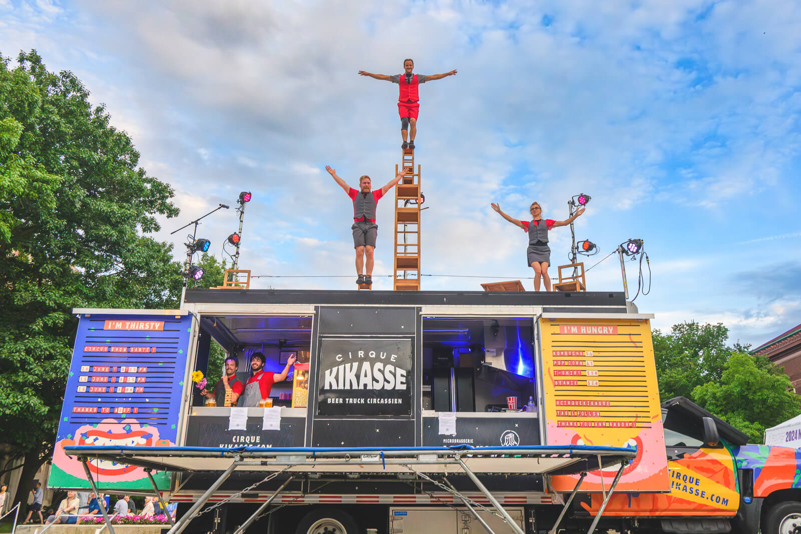 Generate alternate text for this image A vibrant image shows the "Cirque Kikasse" beer truck in Ann Arbor. The truck is colorful and has open panels revealing two individuals inside, seemingly working or serving. On the roof of the truck, three performers are arranged in a pyramid, with one person standing on a ladder held by another, and a third person standing beside them on the roof. The truck has signs that say "I'M THIRSTY" and "I'M HUNGRY," with menus underneath each. The phrase "BEER TRUCK CIRCASSIEN" is also visible. The sky is partly cloudy, and there are trees and buildings in the background.