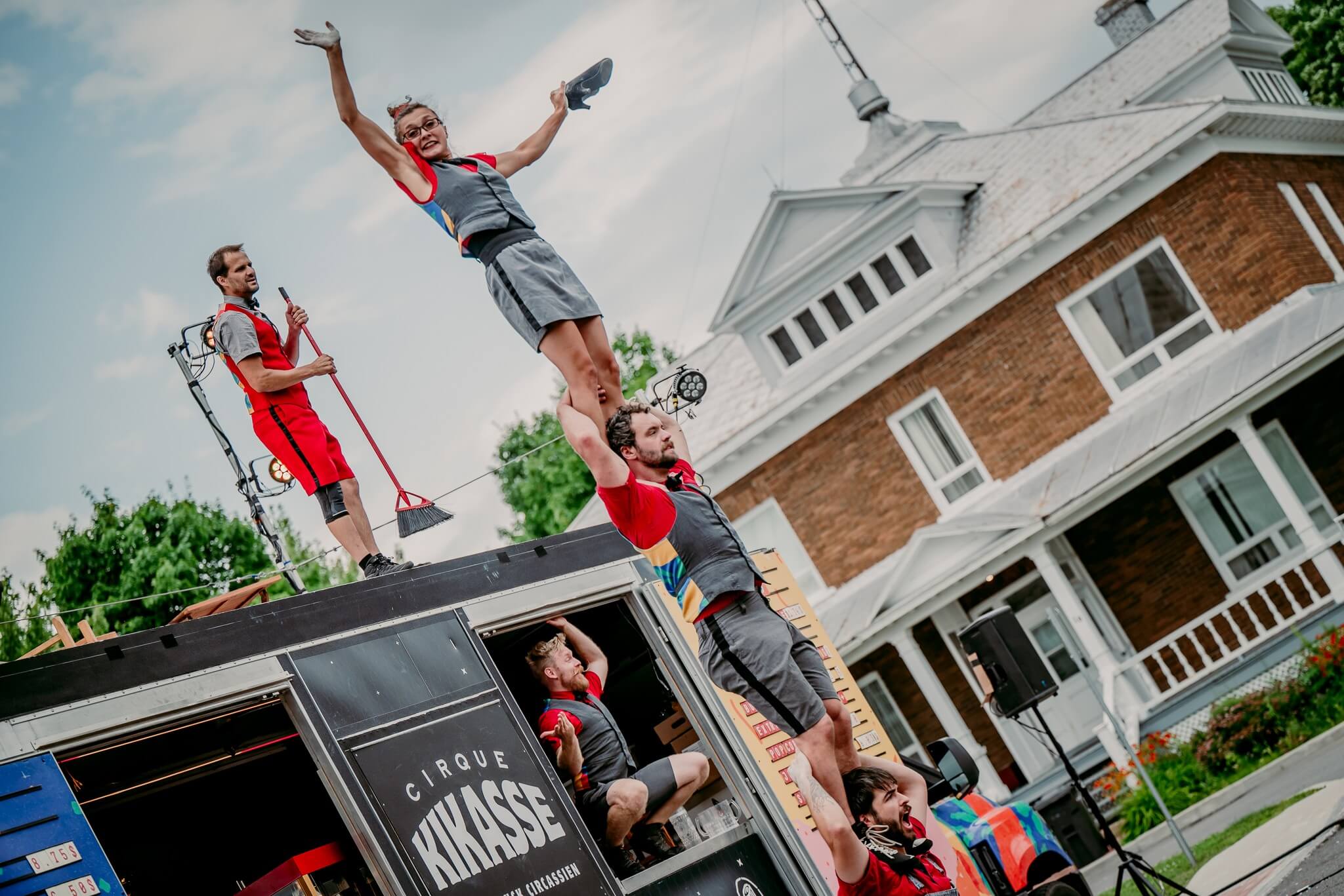 An outdoor shot captures the "Cirque Kikasse" beer truck, which is painted in bright colors. Its side panels are open, revealing two people working inside. On the roof, three performers are arranged in a dynamic pyramid: one person stands on a ladder held by another, while a third person stands beside them on the roof. Signs on the truck read "I'M THIRSTY" and "I'M HUNGRY," with menus displayed below. The text "BEER TRUCK CIRCASSIEN" is also visible. The sky is partly cloudy, and in the background, there are trees and buildings.