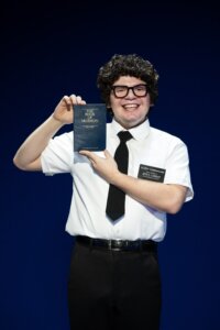 Diego Enrico in THE BOOK OF MORMON North American tour, holding The Book of Mormon, wearing a white shirt and black tie.
Photo by Julieta Cervantes