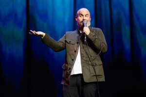Tom Segura on stage, gesturing with a microphone