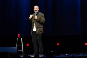 Tom Segura on stage, gesturing with a microphone