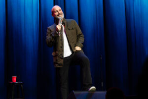 Tom Segura on stage, gesturing with a microphone
