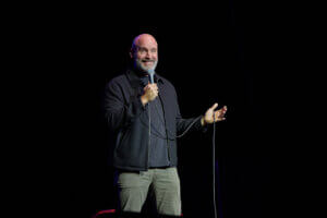 Tom Segura on stage, gesturing with a microphone