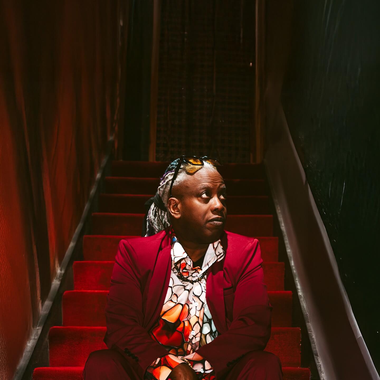Corey Glover of Living Colour wearing a red jacket, sitting on stairs