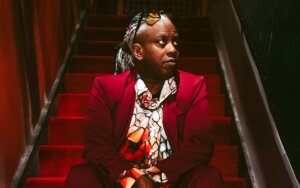 Corey Glover of Living Colour wearing a red suit jacket sitting on red carpeted stairs
