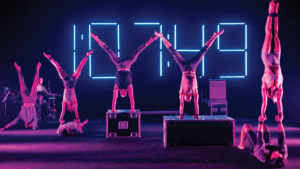 Six performers do handstands on stage in front of a large neon digital clock display, with two standing on boxes and a drummer playing in the background under purple lighting.