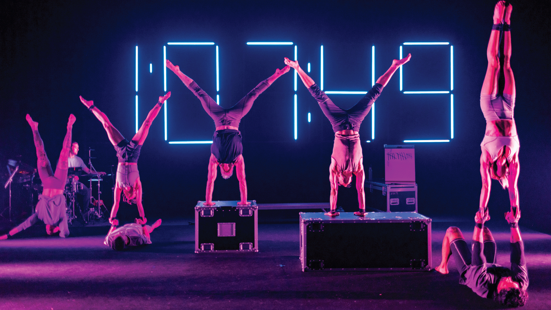 Six performers do handstands on stage in front of a large neon digital clock display, with two standing on boxes and a drummer playing in the background under purple lighting.