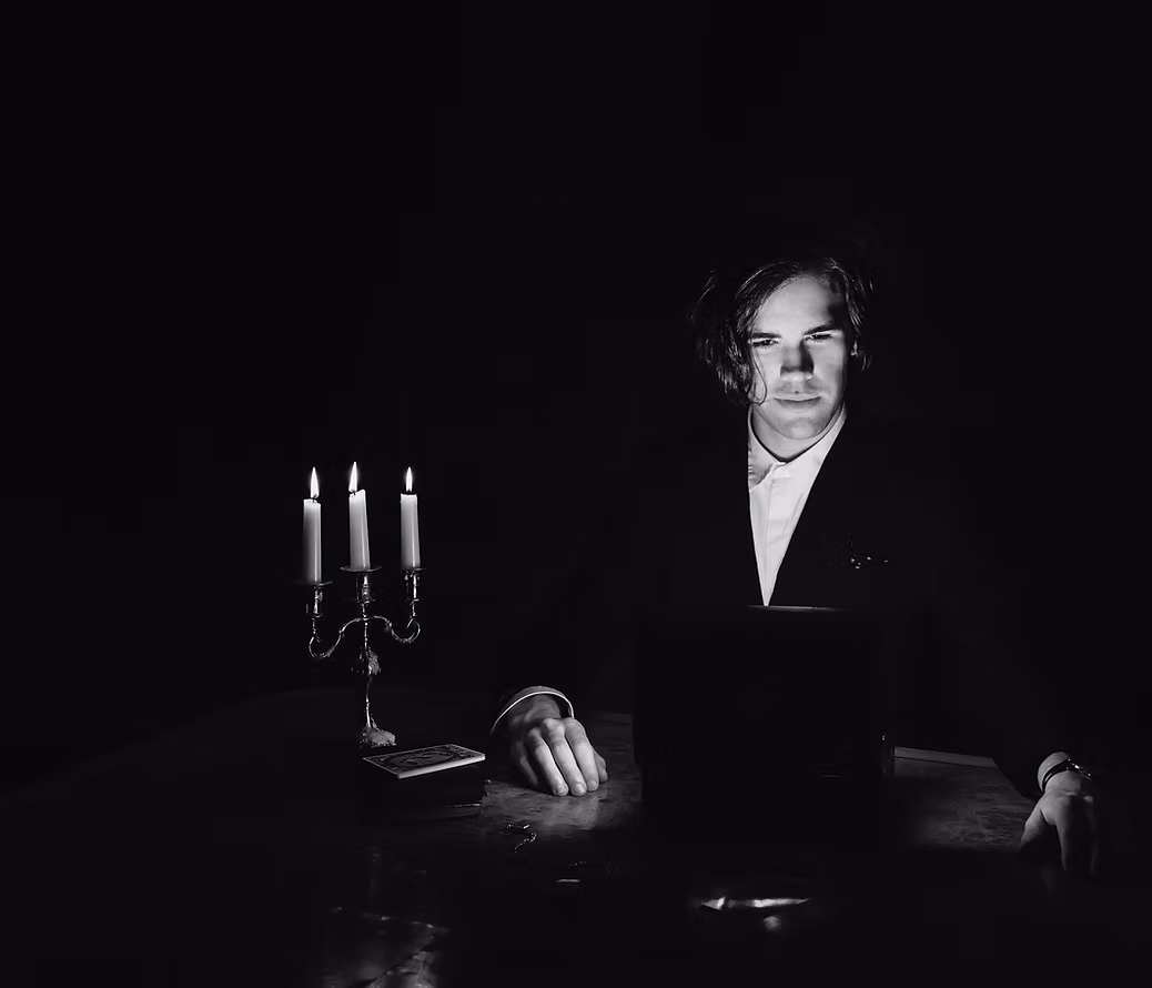 A person in a suit sits at a dimly lit table with a laptop, illuminated by a three-candle candelabra reminiscent of the Illusionist’s Table, creating a dramatic and mysterious atmosphere inspired by Scott Silven.