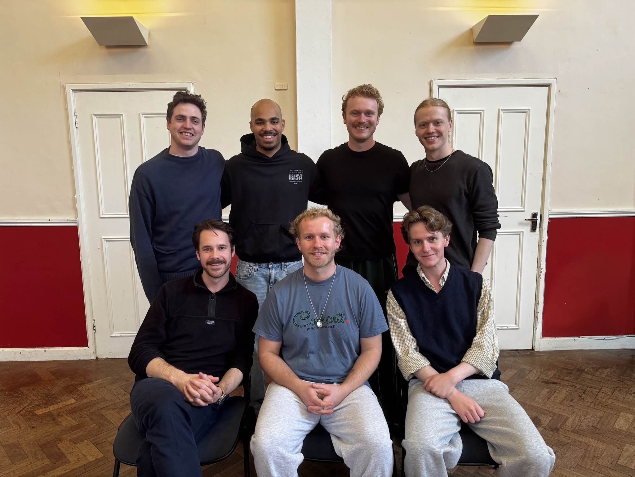 Seven men pose together in a brightly lit room with cream walls and red paneling. Three are seated in front, while four stand behind them. They are all smiling and dressed casually, perhaps cast members from a Shakespeare play like Othello by William Shakespeare.