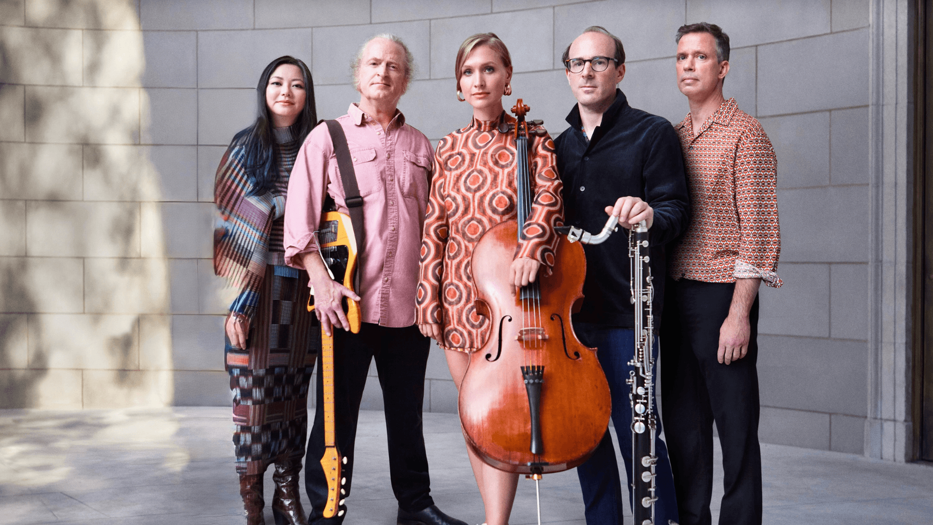 Five musicians from the Bang On A Can All-Stars music ensemble stand together holding their instruments, including an electric guitar, cello, and woodwind, against a light stone wall. They wear patterned, colorful clothing and face the camera as a group.