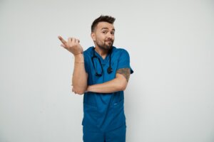 Nurse Blake, dressed in blue medical scrubs with a stethoscope around his neck, stands against a plain white background, arms crossed as he gestures playfully to the side with a questioning expression.