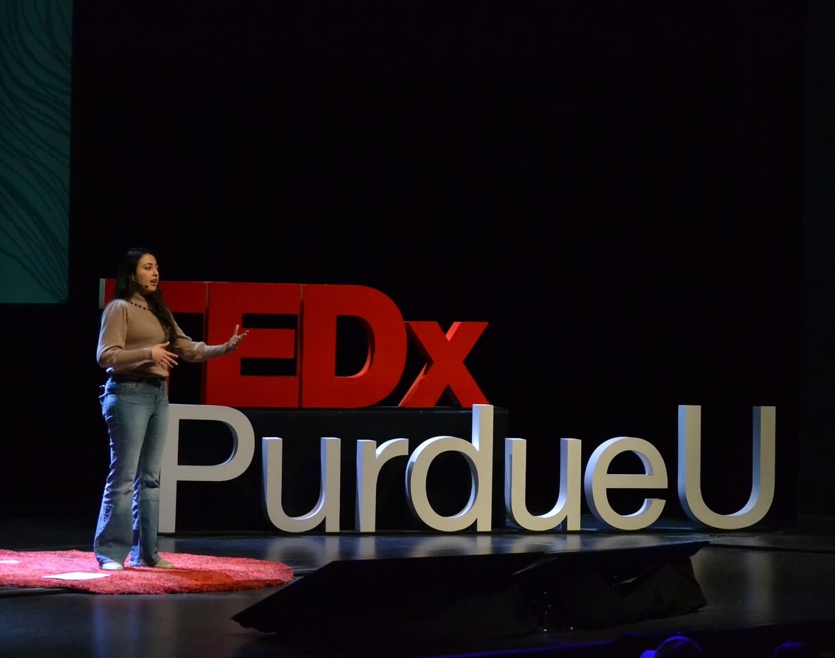 A woman speaks on stage at TEDxPurdueU in front of large red and white letters, standing on a red circular carpet with a dark background, shedding light on the Unseen.