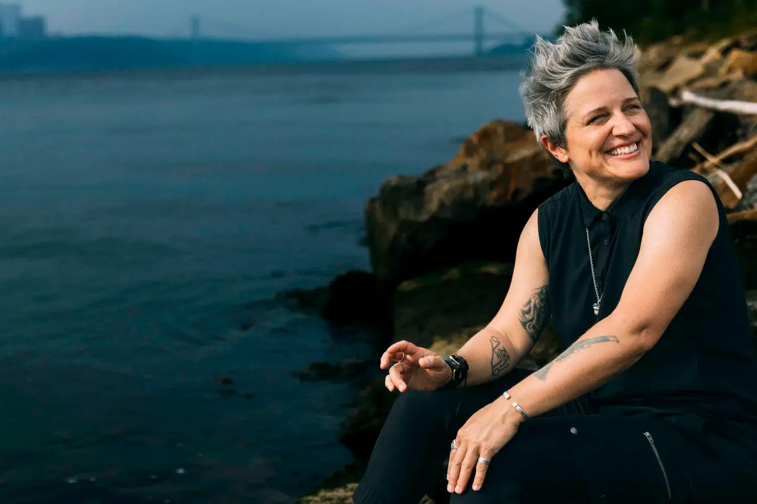 A person with short gray hair and tattoos, wearing a sleeveless black shirt, sits on rocks by a body of water, smiling and looking to the side with a bridge visible in the background—reminiscent of jazz band leader Allison Miller of Boom Tic Boom.