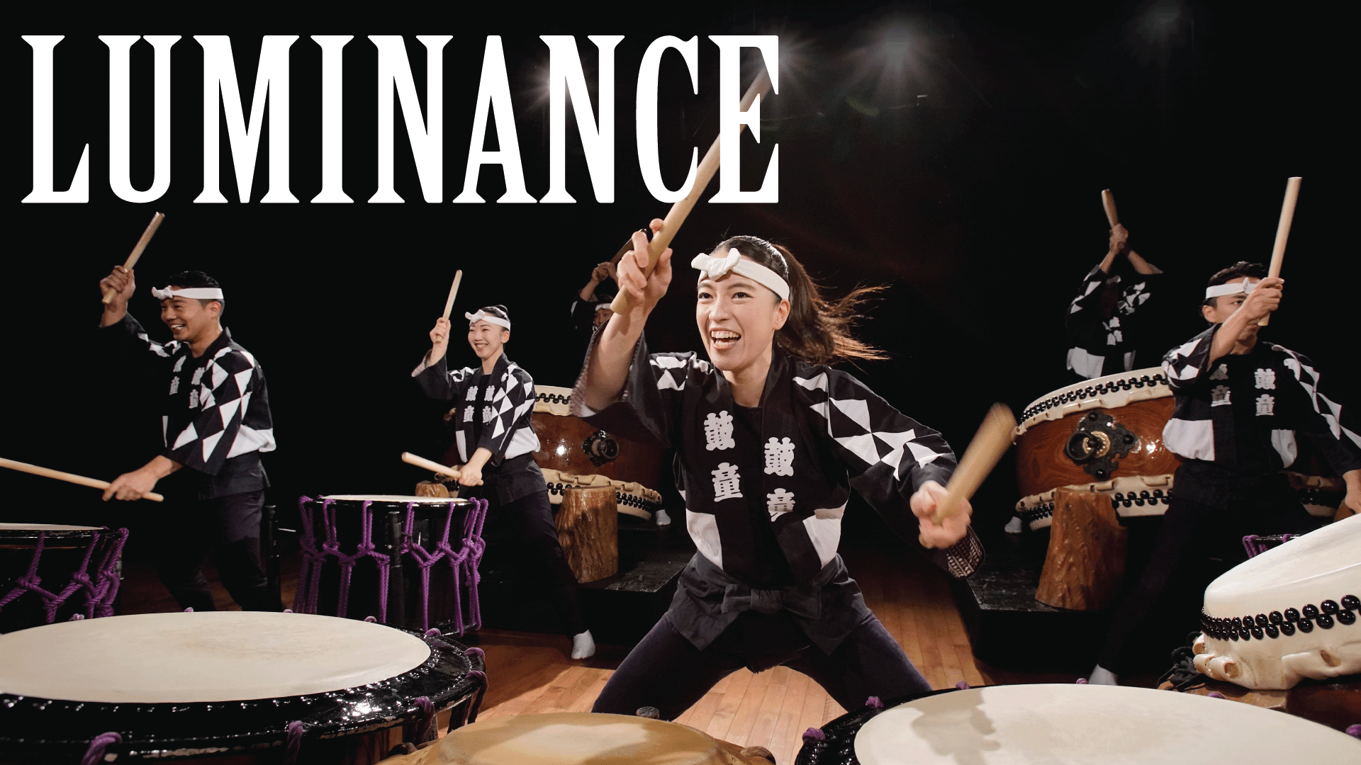 A group of enthusiastic taiko drummers perform on stage, smiling and wearing matching black and white outfits. The word LUMINANCE appears in bold letters at the top left of the image.