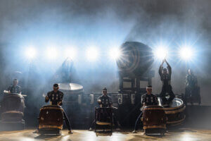 Six traditional KODO Japanese taiko drummers perform on stage with large drums, illuminated by bright luminance behind them, creating dramatic silhouettes and a smoky atmosphere.