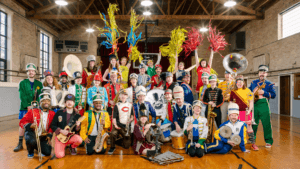 A diverse group of people in colorful, mismatched marching band uniforms pose with various instruments in a gymnasium. Some hold brass and woodwind instruments, while others wave bright pom-poms. They are smiling and playful.