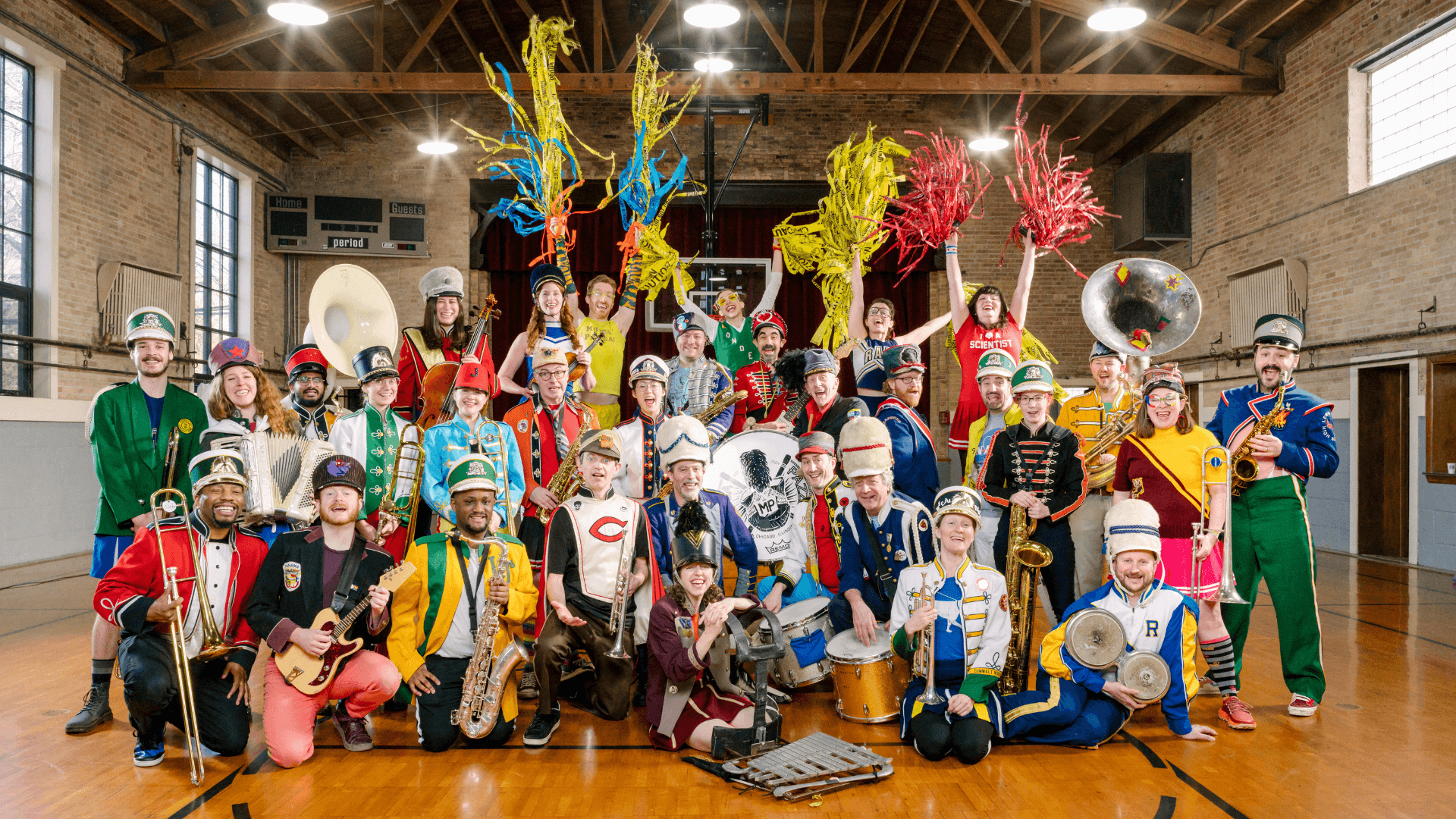 A diverse group of people in colorful, mismatched marching band uniforms pose with various instruments in a gymnasium. Some hold brass and woodwind instruments, while others wave bright pom-poms. They are smiling and playful.
