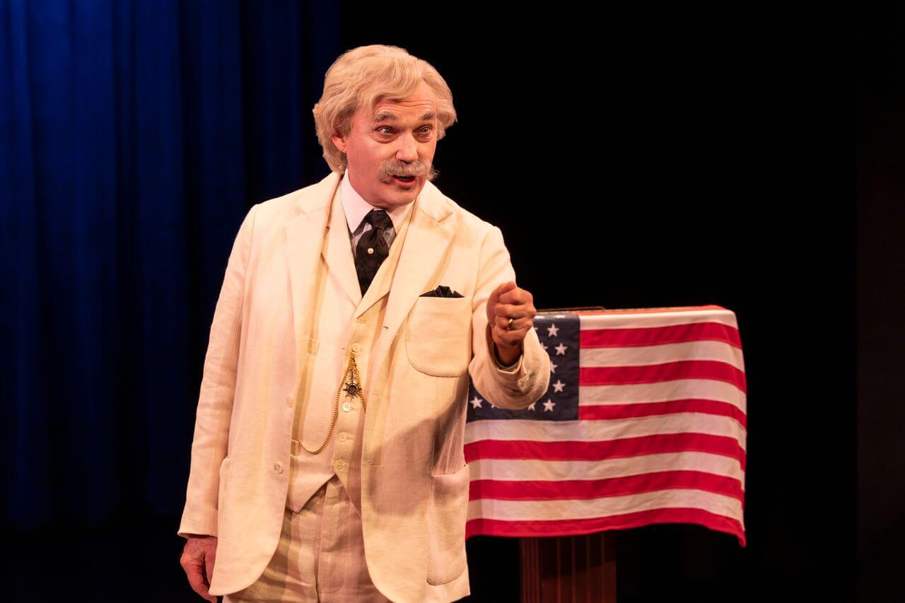 An older man in a white suit gestures while speaking on stage next to a podium draped with an American flag, reminiscent of Mark Twain Tonight. The background is dark with blue stage lighting.