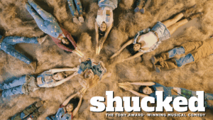 A group of people dressed in country attire lies in a circle on straw, holding hands and corn cobs in the center. The text reads: shucked: The Tony Award-winning musical comedy.
