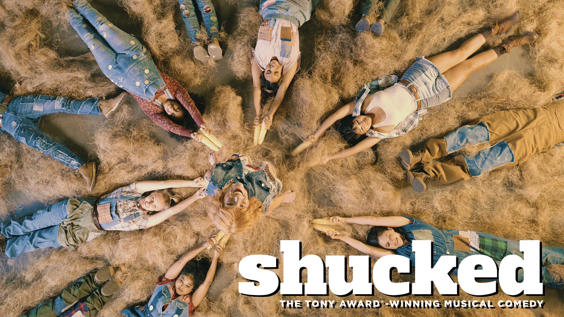 A group of people dressed in country attire lies in a circle on straw, holding hands and corn cobs in the center. The text reads: shucked: The Tony Award-winning musical comedy.