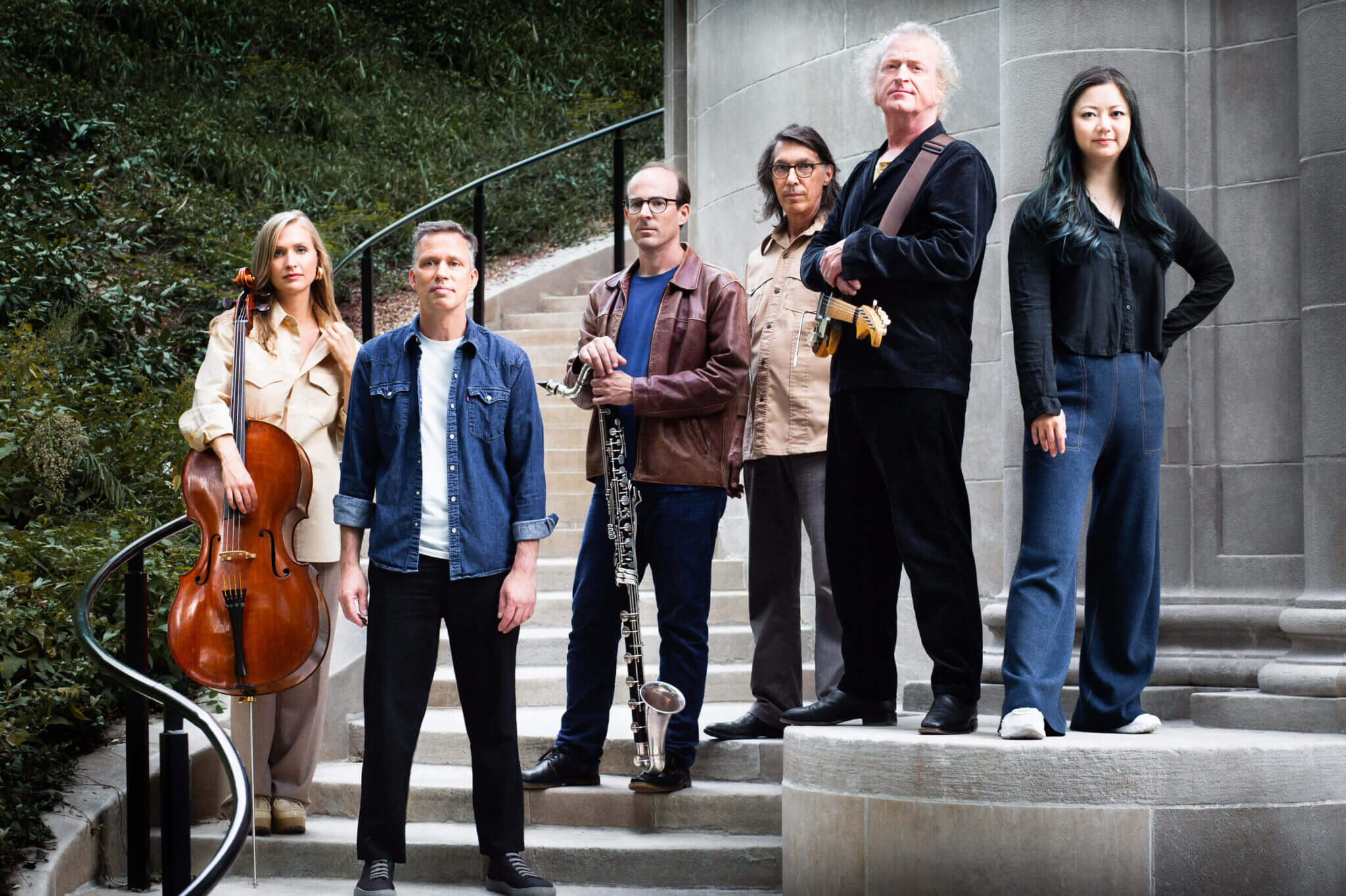 Six people, the Bang On A Can All-Stars, stand on outdoor stone steps. One holds a cello, another a clarinet, and a third an electric guitar. They pose in casual and semi-formal clothing by a curved metal railing, with greenery behind them.