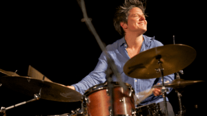 Allison Miller, sporting short hair and a blue shirt, smiles while playing the drum set on stage for Boom Tic Boom, drumsticks in hand and a microphone stand in the foreground.