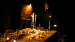 A man in a suit stands in dim lighting beside a candle-lit dining table set for several people, an easel and bottle in the background, evoking the mysterious and moody atmosphere of Scott Silven’s Illusionist’s Table immersive experience.