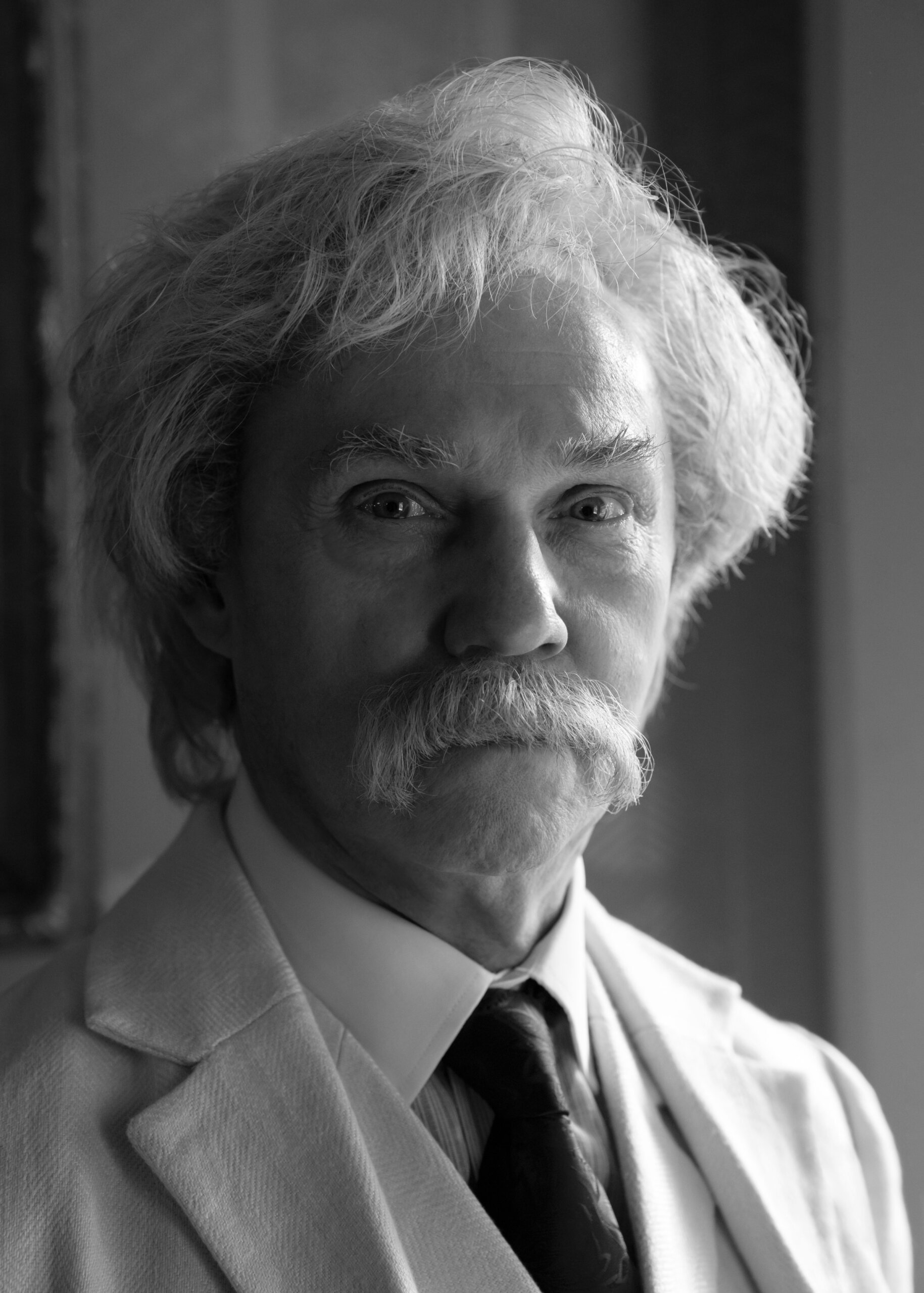 An older man in a white suit gestures while speaking on stage next to a podium draped with an American flag, reminiscent of Mark Twain Tonight. The background is dark with blue stage lighting.