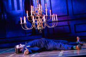 A person in a suit lies flat on their back on a stage floor beneath a large, lit chandelier, with dramatic blue lighting and dark panelled walls in the background.