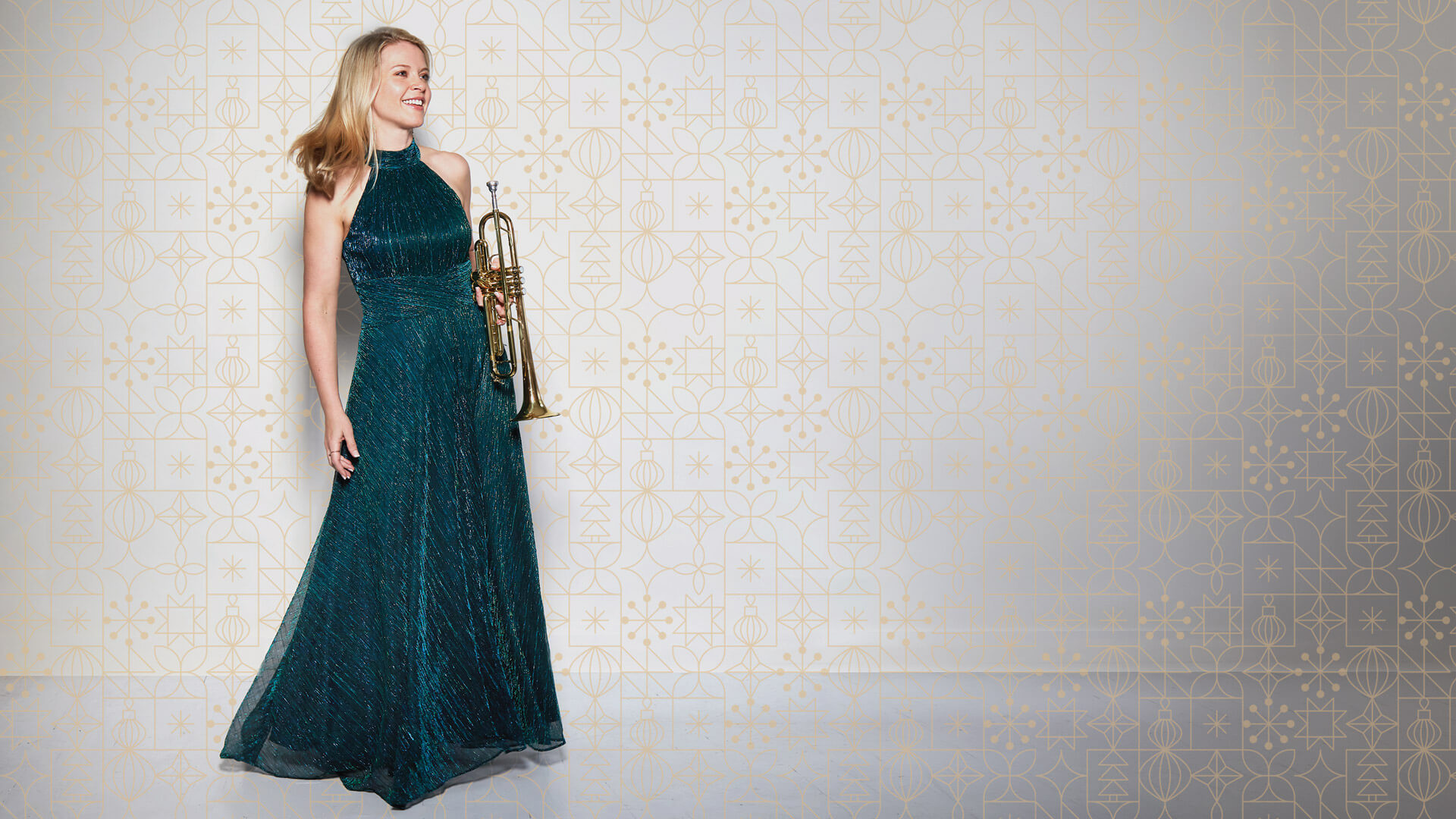 Bria Skonberg wearing a sleeveless, floor-length teal gown stands holding a trumpet. She is smiling and looking to the side, reminiscent of the festive spirit in Bria Skonberg's Jingle Bell Swing, with a patterned beige and gold background behind her.