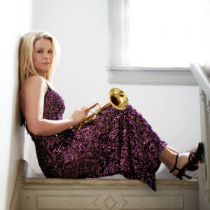 A woman in a purple sequined dress sits on stairs, leaning against a wall and holding a trumpet. Sunlight comes through a window beside her.