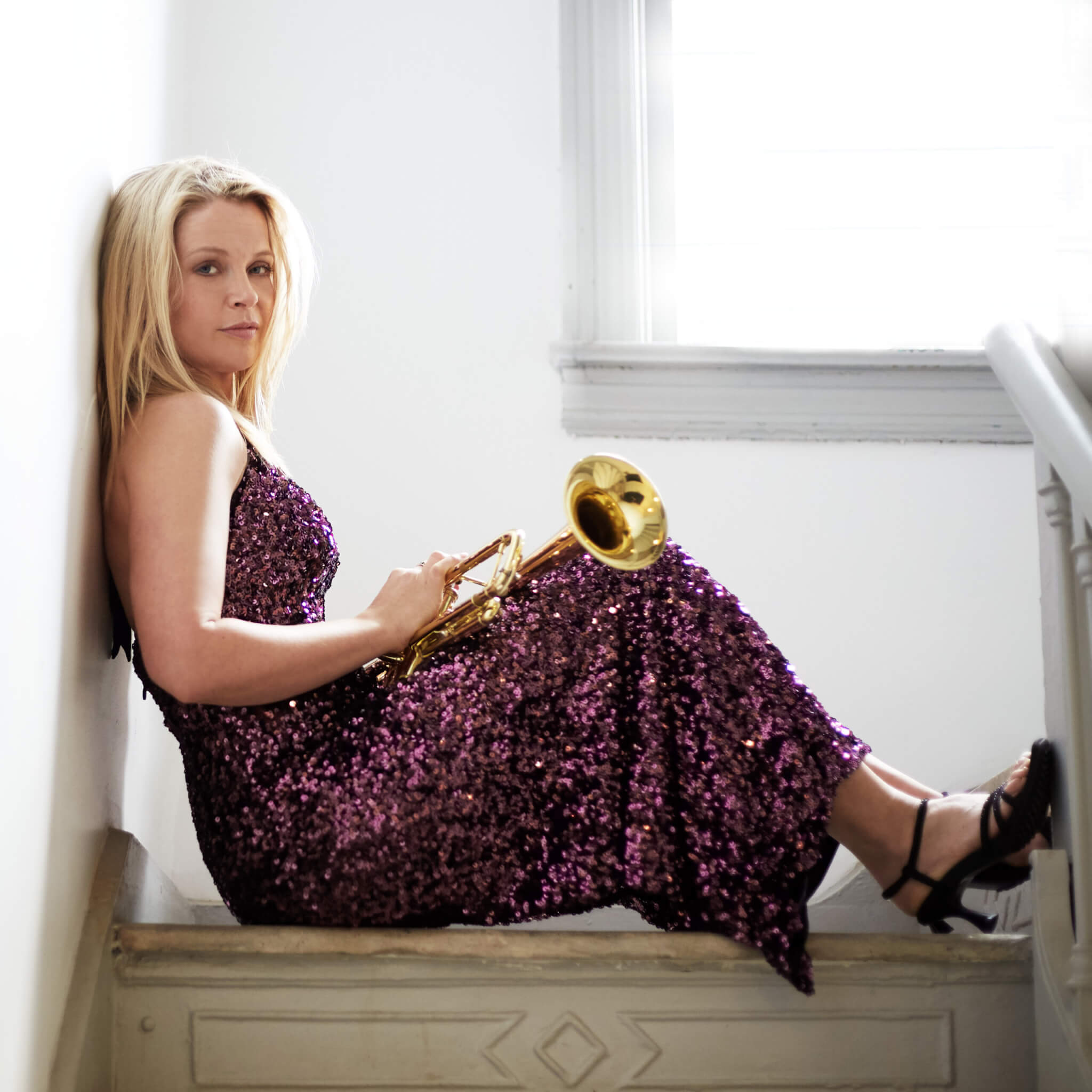 A woman in a purple sequined dress sits on stairs, leaning against a wall and holding a trumpet. Sunlight comes through a window beside her.