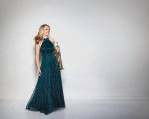 A woman in a long, elegant dark green dress stands against a plain white wall, smiling and holding a brass trumpet by her side.