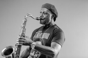 Black and white photo of a man in a beret and T-shirt playing a saxophone, focused on his performance. Captured against a plain background, this image celebrates the spirit of the 30th Annual West Lafayette Global Fest.