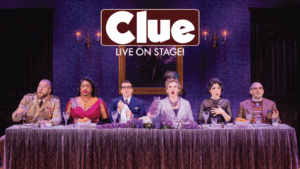 Six actors in colorful costumes, sit at a long dining table on stage, each with dramatic expressions. The backdrop is purple, and a “Clue LIVE ON STAGE!” sign hangs above them.