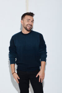 A man with short brown hair and a beard, wearing a dark blue sweater and black pants, stands smiling against a plain white background.