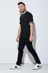 A man with short hair and a beard, wearing a black t-shirt, black pants, and white sneakers with black accents, walks confidently while smiling against a plain white background.