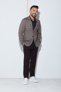 A man with short hair and a beard stands smiling against a plain white wall, wearing a checked blazer, dark pants, a dark shirt, and white sneakers. His hands are in his pockets and he leans slightly to one side.