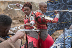 Two women in red and silver armor stand back-to-back, holding circular weapons and spears, ready for battle. Inspired by Marvel Studios' Black Panther Live In Concert, they face off against shielded opponents on a rocky outdoor backdrop.